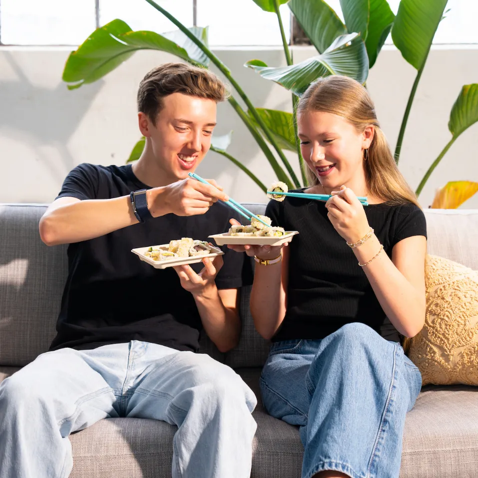 Two friends sitting on a couch sharing Happy Rice sushi with teal chopsticks