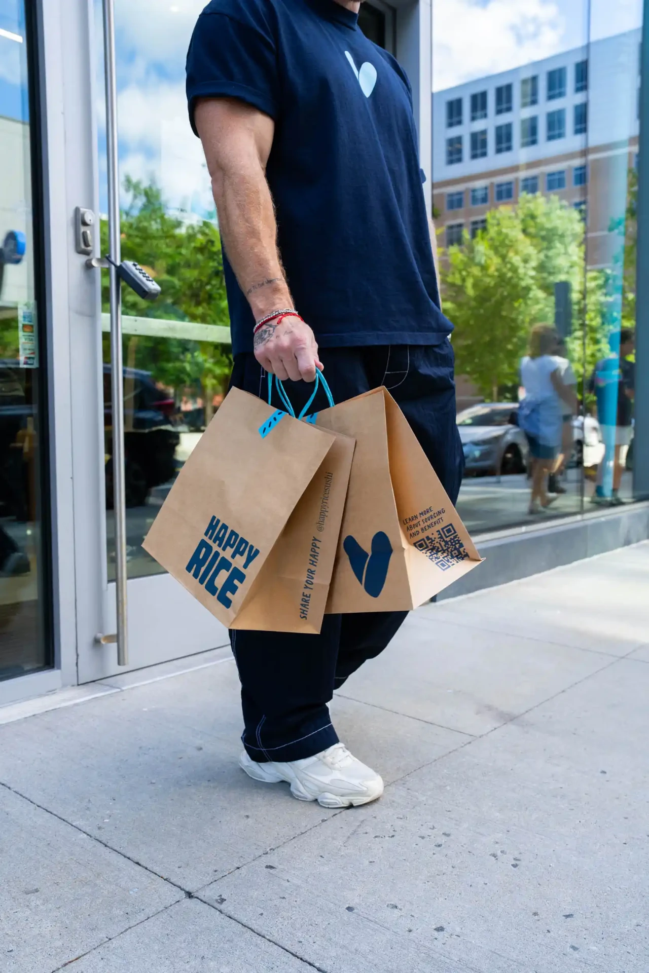 Person in a navy Happy Rice T-shirt carrying two Happy Rice takeout bags outside the Boston Seaport restaurant