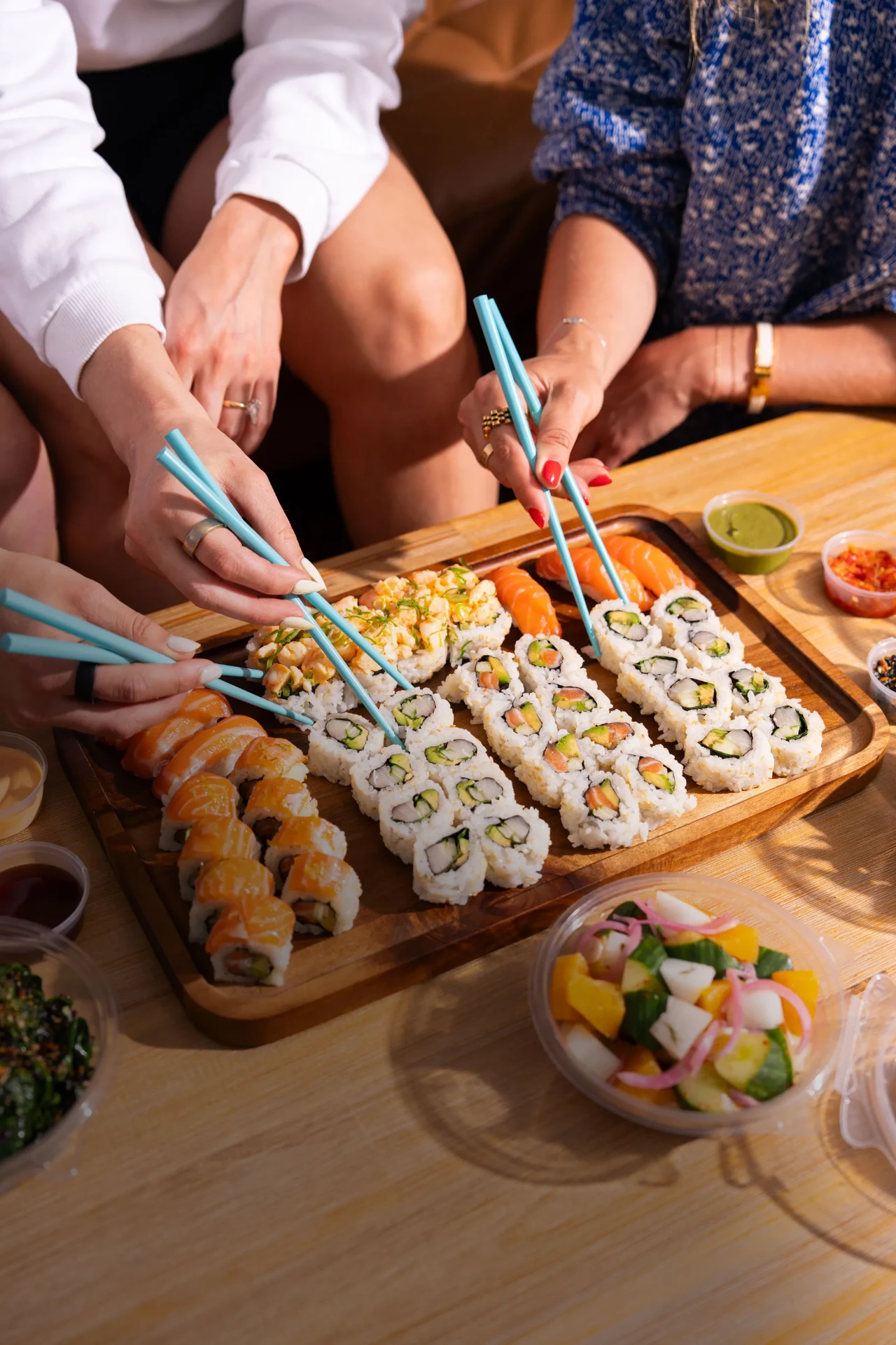 Friends sharing a wooden board of Happy Rice sushi rolls with teal chopsticks