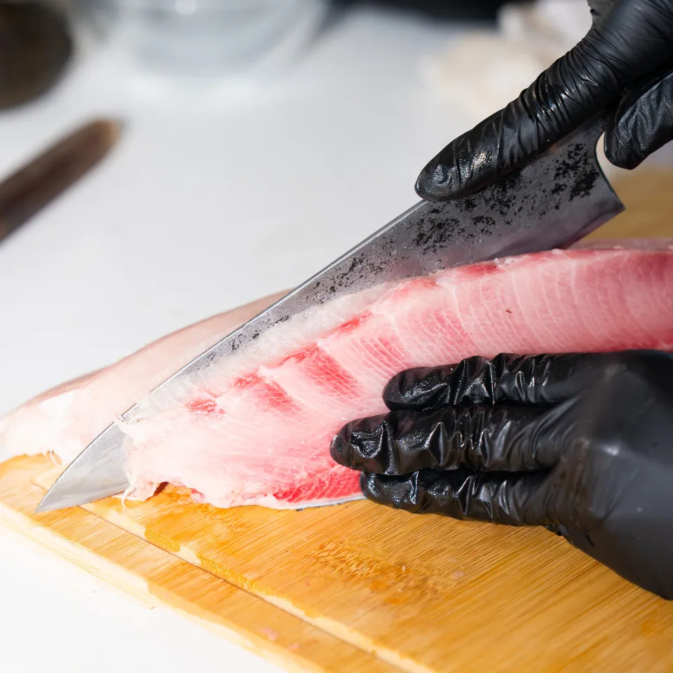 Gloved hands slicing a fillet of fish with a long knife on a wooden board
