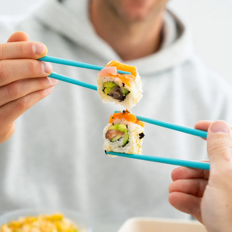 Two hands holding pieces of Happy Rice sushi with teal chopsticks