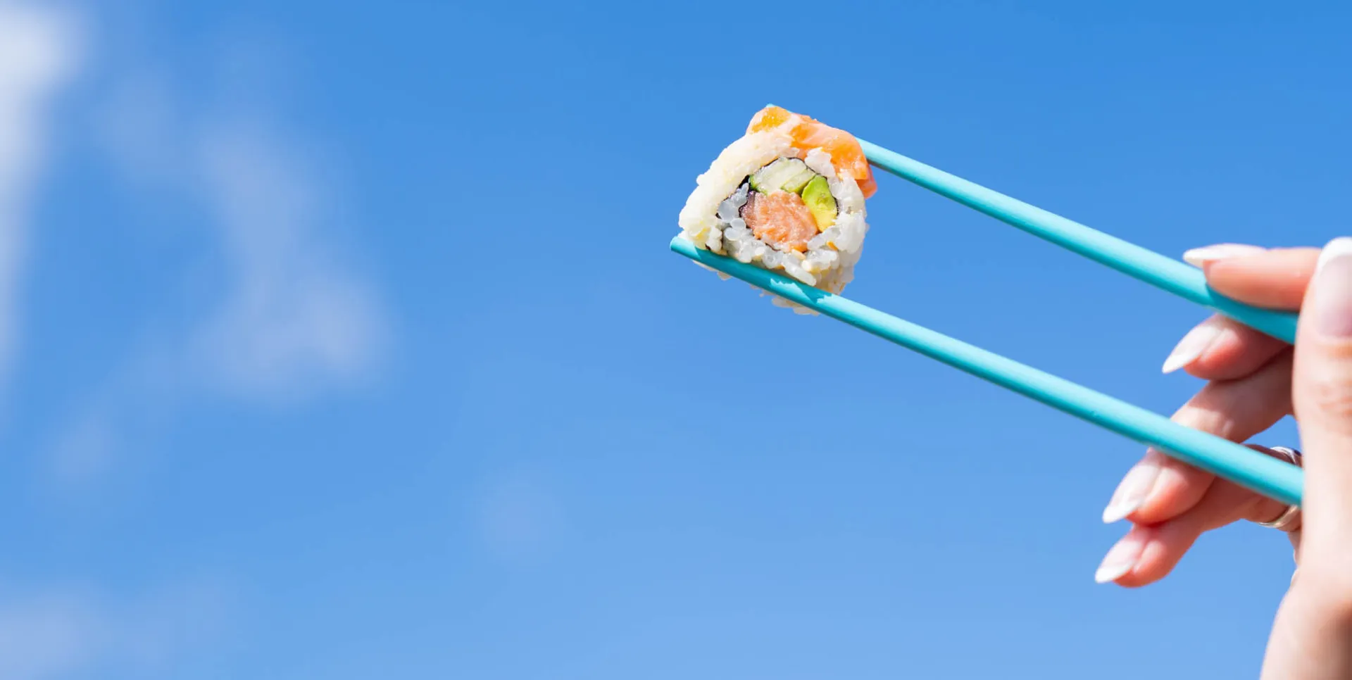 Hand holding chopsticks with salmon sushi against a blue sky