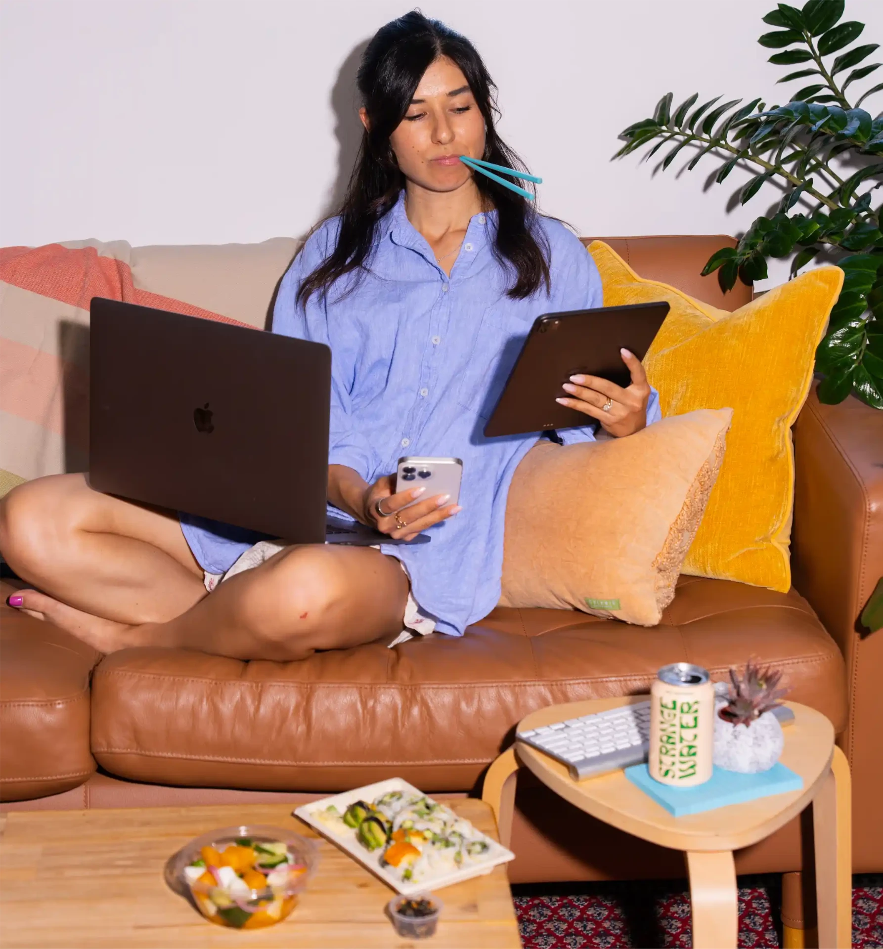 Woman on a leather couch with a laptop, tablet and phone, teal chopsticks in her mouth, with Happy Rice sushi and a salad on the coffee table