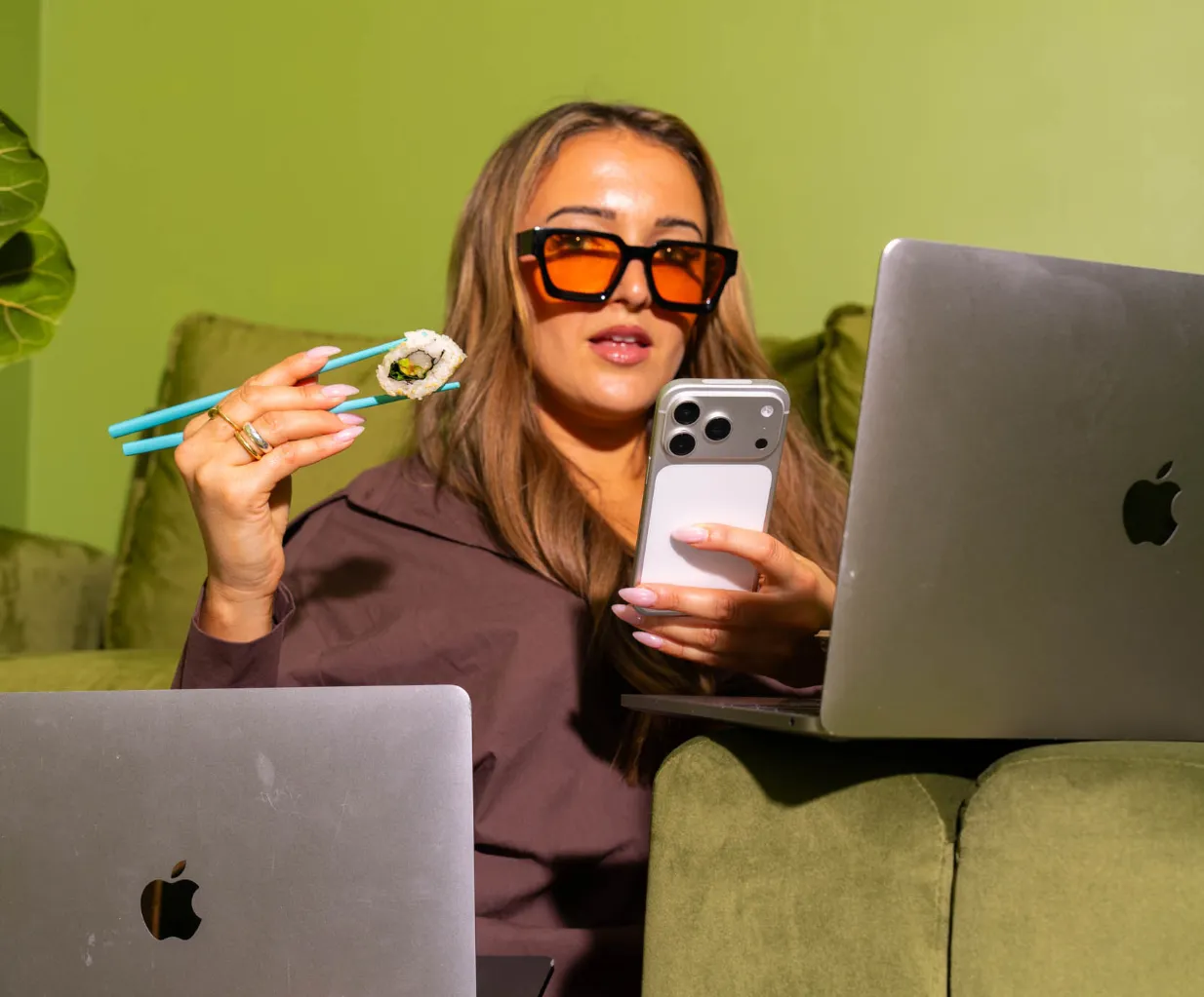 Woman in orange sunglasses eating a sushi roll with teal chopsticks while checking her phone between two laptops