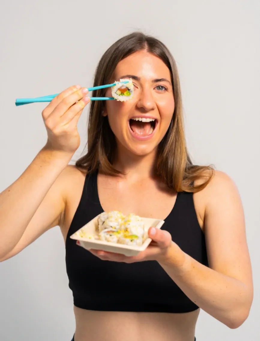 Smiling Happy Rice team member holding a tray of sushi rolls and a roll up to her eye with teal chopsticks
