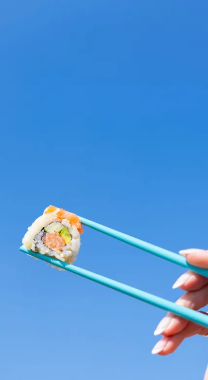 Hand holding chopsticks with salmon sushi against a blue sky