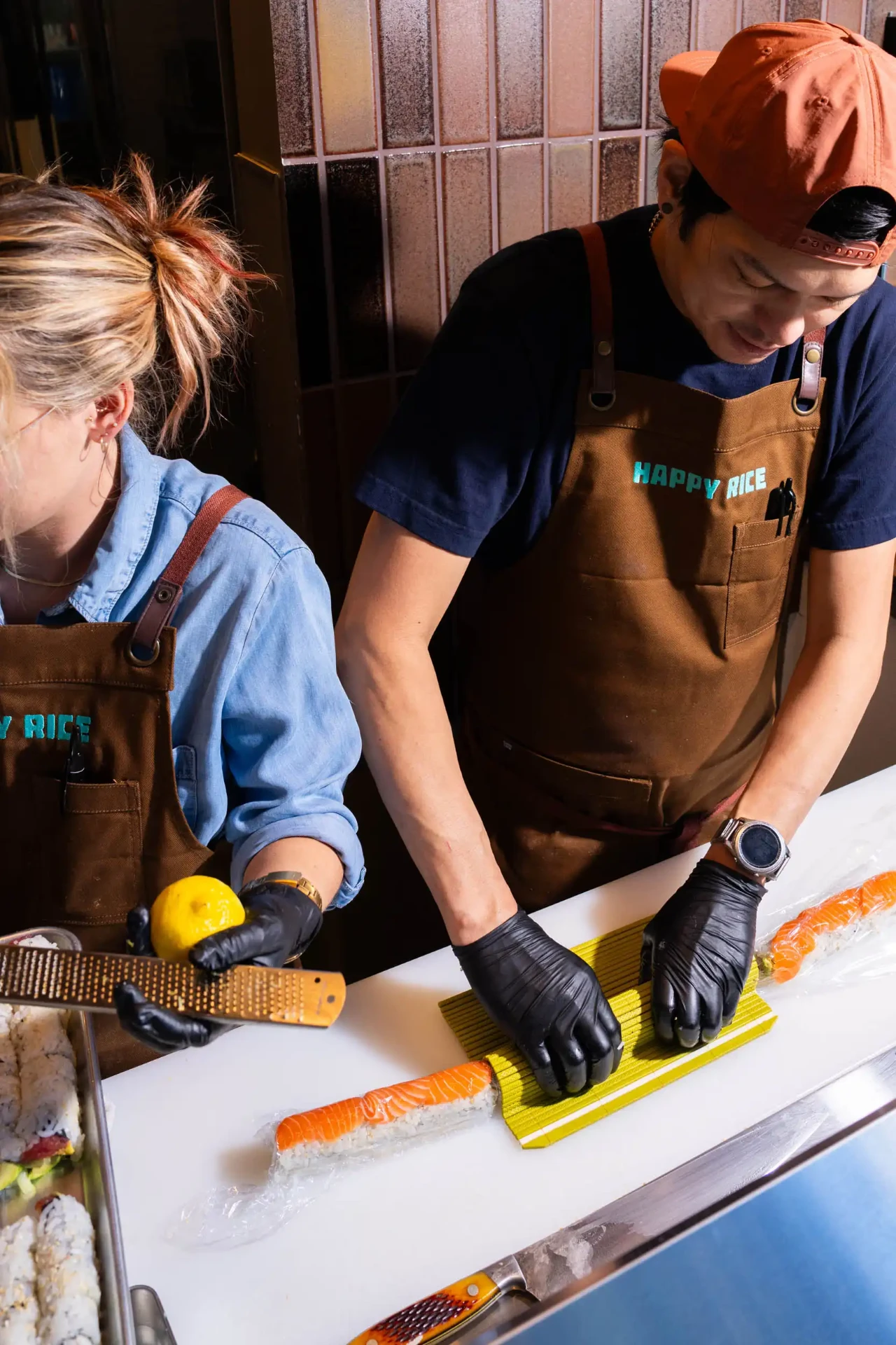 Two Happy Rice chefs in branded aprons rolling salmon sushi and zesting lemon at the counter